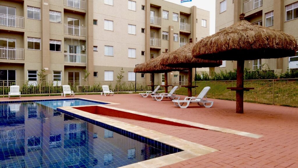 Área de lazer do empreendimento Quali Bonfim Paulista com piscina revestida de azulejos azuis, espreguiçadeiras brancas e quiosques com cobertura de palha em frente aos blocos residenciais.